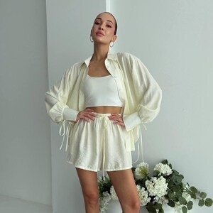 Könnte beinhalten: Ein cremefarbenes zweiteiliges Set bestehend aus einem langärmeligen Hemd mit Knopfleiste und Bindedetails an den Manschetten sowie passenden Shorts mit hoher Taille und elastischem Kordelzug, getragen über einem weißen Top mit weißen Pantoletten.