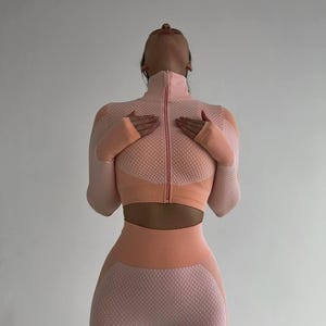 Op de afbeelding: Een tweedelige activewear set in perzik- en lichtroze tinten. De set bestaat uit een crop top met lange mouwen en ritssluiting en een legging met hoge taille. De stof heeft een getextureerd, gestippeld patroon. Het model draagt de outfit.