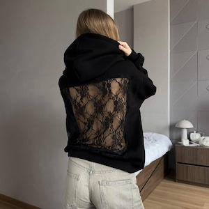 Op de afbeelding: Zwarte hoodie met een kanten achterpaneel. De hoodie heeft een capuchon met trekkoord en lange mouwen. Het kanten paneel heeft een donker, ingewikkeld ontwerp. De persoon draagt een lichtgekleurde jeans.