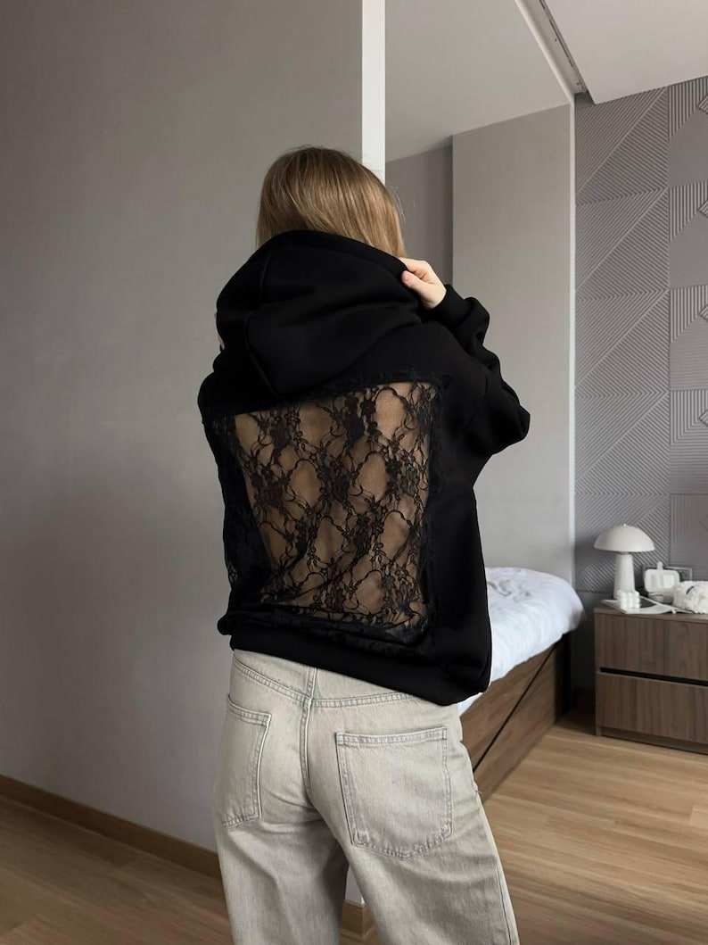 Op de afbeelding: Zwarte hoodie met een kanten achterpaneel. De hoodie heeft een capuchon met trekkoord en lange mouwen. Het kanten paneel heeft een donker, ingewikkeld ontwerp. De persoon draagt een lichtgekleurde jeans.