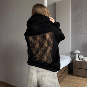 Op de afbeelding: Zwarte hoodie met een kanten achterpaneel. De hoodie heeft een capuchon met trekkoord en lange mouwen. Het kanten paneel heeft een donker, ingewikkeld patroon. De persoon draagt een lichtgekleurde jeans.