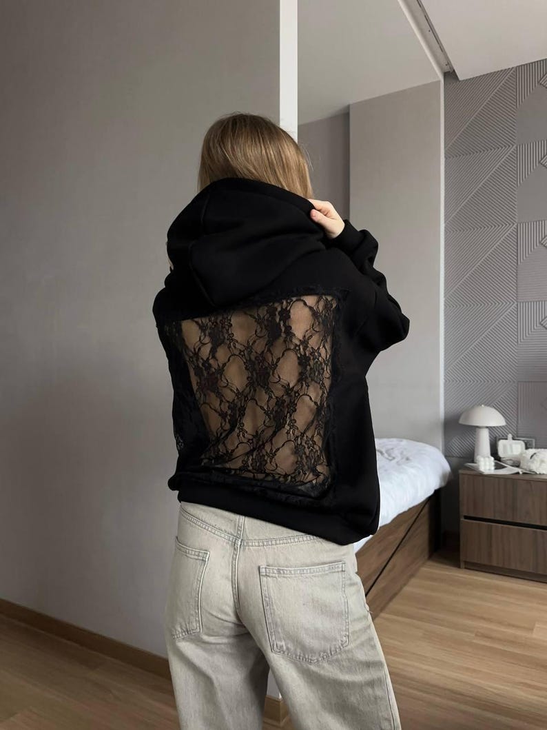Op de afbeelding: Zwarte hoodie met een kanten achterpaneel. De hoodie heeft een capuchon met trekkoord en lange mouwen. Het kanten paneel heeft een donker, ingewikkeld patroon. De persoon draagt een lichtgekleurde jeans.