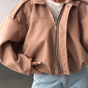Op de afbeelding: Een beige jas met lange mouwen, een omgeslagen kraag, een ritssluiting over de hele lengte en een drukknoopsluiting. De jas heeft gestructureerde schouders, een korte zoom en een gladde, effen stoffen afwerking, gedragen over een wit shirt.