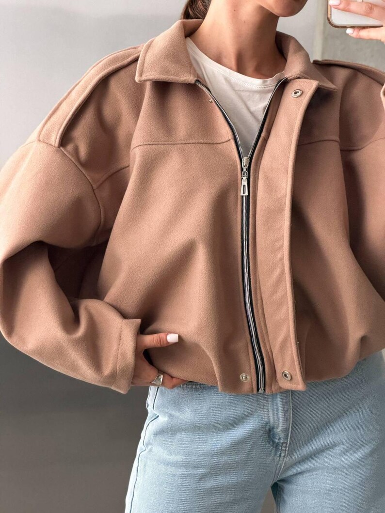 Op de afbeelding: Een beige jas met lange mouwen, een omgeslagen kraag, een ritssluiting over de hele lengte en een drukknoopsluiting. De jas heeft gestructureerde schouders, een korte zoom en een gladde, effen stoffen afwerking, gedragen over een wit shirt.