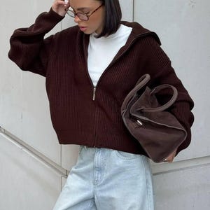 May include: A dark brown, zip-up knitted sweater with a hood, worn over a white turtleneck. The outfit includes light blue jeans and a matching brown handbag. The sweater has a ribbed texture.