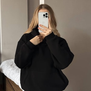 Op de afbeelding: Een zwarte hoodie met een voorzak. De hoodie is gemaakt van een zacht materiaal en heeft een relaxte pasvorm. De persoon op de foto draagt een lichtgekleurde broek.