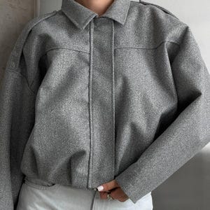 Op de afbeelding: Een grijze jas met kraag en een textuurafwerking, voorzien van lange mouwen, een knoopsluiting aan de voorzijde en een korte zoom. De jas heeft een gestructureerde kraag en schouderdetails, gecombineerd met een lichtgekleurde spijkerbroek.