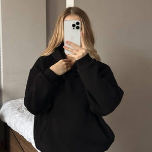 Op de afbeelding: Een zwarte hoodie met een voorzak. De hoodie is gemaakt van een zacht materiaal en heeft een relaxte pasvorm. De persoon op de foto draagt een lichtgekleurde broek.