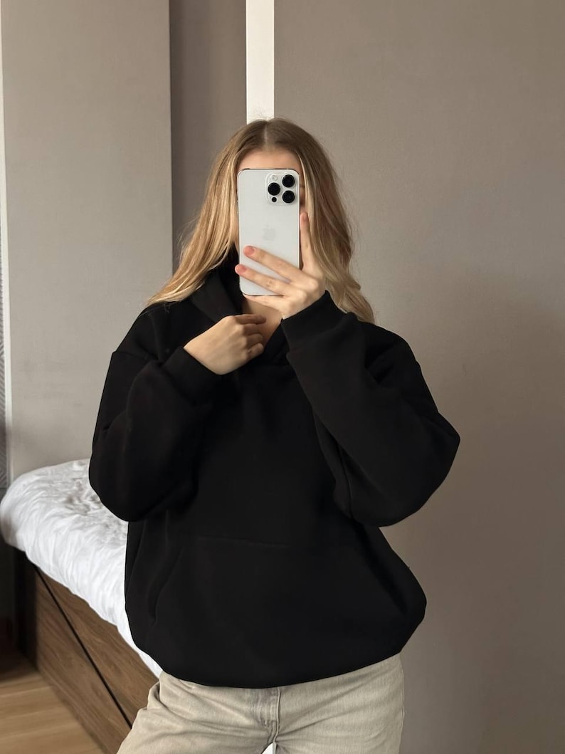 Op de afbeelding: Een zwarte hoodie met een voorzak. De hoodie is gemaakt van een zacht materiaal en heeft een relaxte pasvorm. De persoon op de foto draagt een lichtgekleurde broek.