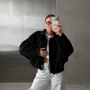 Op de afbeelding: Een zwart kort jasje met een kraag en ritssluiting aan de voorzijde, gedragen over een wit t-shirt. De outfit bevat een lichtblauwe spijkerbroek met een grote, versleten scheur op het bovenbeen. Afgemaakt met een bril met donker montuur.