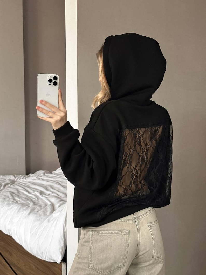 Op de afbeelding: Zwarte hoodie met een kanten achterpaneel. De hoodie heeft een capuchon met trekkoord en lange mouwen. Het model draagt een lichtgekleurde jeans en houdt een smartphone vast.
