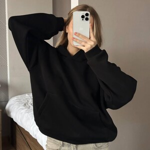 Op de afbeelding: Een zwarte hoodie met een kangoeroezak. De hoodie heeft lange mouwen en een capuchon. De persoon op de foto draagt een lichtgekleurde jeans. De achtergrond is neutraal.
