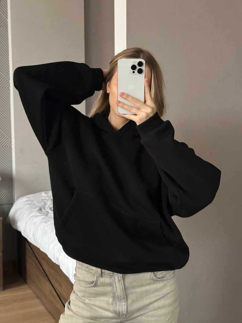 Op de afbeelding: Een zwarte hoodie met een kangoeroezak. De hoodie heeft lange mouwen en een capuchon. De persoon op de foto draagt een lichtgekleurde jeans. De achtergrond is neutraal.