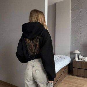 Op de afbeelding: Zwarte cropped hoodie met een kanten achterpaneel. De hoodie heeft een capuchon met trekkoord en lange mouwen. De persoon draagt een lichtgeleurde jeans en draagt een kleine zwarte tas met een kettingriem. De achtergrond is een kamer met neutrale tinten.