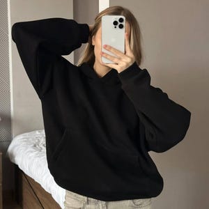 Op de afbeelding: Een zwarte hoodie met een kangoeroezak. De hoodie heeft lange mouwen en een capuchon. De persoon draagt een lichtgekleurde jeans. De achtergrond omvat een bed en een muur.