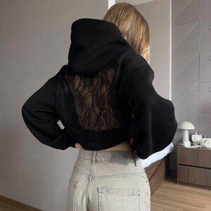 Op de afbeelding: Zwarte cropped hoodie met een kanten achterpaneel. De hoodie heeft een capuchon met trekkoord en lange mouwen. Het model draagt een lichtgekleurde jeans. De algehele stijl is casual en modieus.