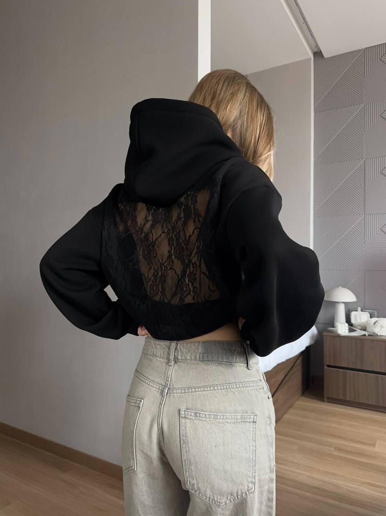Op de afbeelding: Zwarte cropped hoodie met een kanten achterpaneel. De hoodie heeft een capuchon met trekkoord en lange mouwen. Het model draagt een lichtgekleurde jeans. De algehele stijl is casual en modieus.