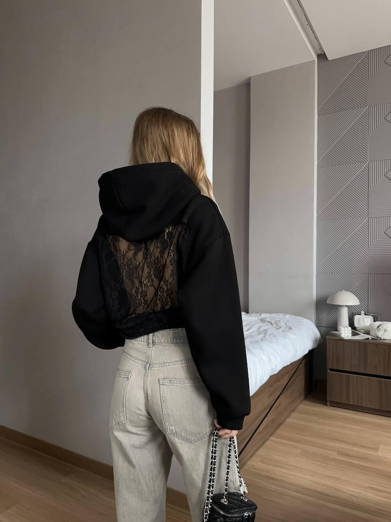 Op de afbeelding: Zwarte cropped hoodie met een kanten achterpaneel. De hoodie heeft een capuchon met trekkoord en lange mouwen. De persoon draagt een lichtgekleurde jeans en draagt een kleine zwarte gewatteerde tas met een kettingriem.