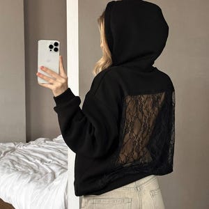 Op de afbeelding: Zwarte hoodie met een kanten achterpaneel. De hoodie heeft een capuchon met trekkoord en lange mouwen. Het model draagt een lichtgekleurde jeans en houdt een smartphone vast.