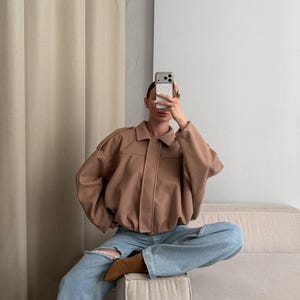 Op de afbeelding: Een beige jas met kraag, lange mouwen en een knoopsluiting, gecombineerd met een lichtblauwe spijkerbroek met een scheur bij de knie en bruine laarzen met een spitse neus. De outfit wordt gedragen met een smartphone voor het gezicht.