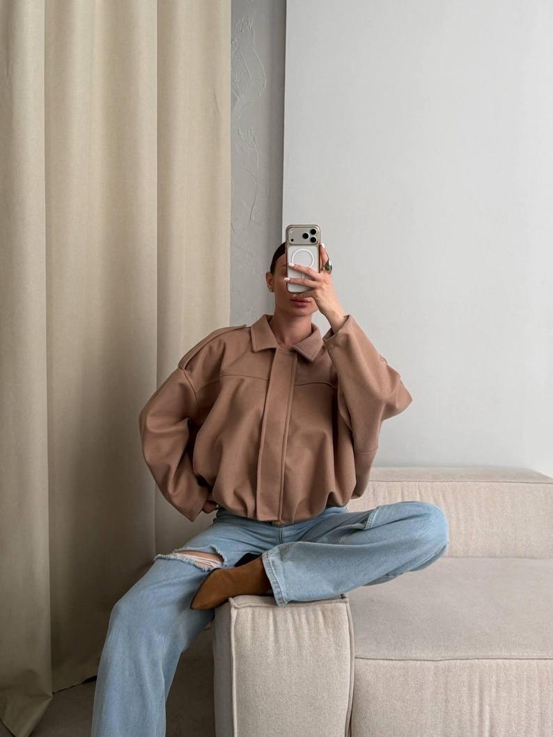 Op de afbeelding: Een beige jas met kraag, lange mouwen en een knoopsluiting, gecombineerd met een lichtblauwe spijkerbroek met een scheur bij de knie en bruine laarzen met een spitse neus. De outfit wordt gedragen met een smartphone voor het gezicht.