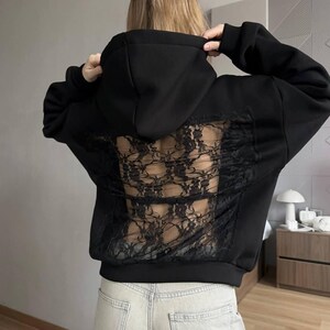 Op de afbeelding: Zwarte hoodie met een kanten achterpaneel. De hoodie heeft een capuchon met trekkoord en lange mouwen. De achterkant is gemaakt van zwart kant, wat een uniek ontwerp cre&euml;ert.