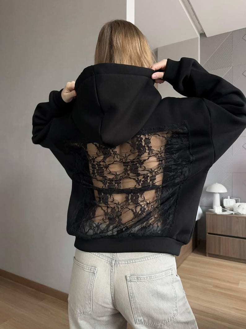Op de afbeelding: Zwarte hoodie met een kanten achterpaneel. De hoodie heeft een capuchon met trekkoord en lange mouwen. De achterkant is gemaakt van zwart kant, wat een uniek ontwerp cre&euml;ert.