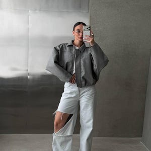 Op de afbeelding: Een grijs kort jasje met kraag en lange mouwen gedragen over een wit shirt, gecombineerd met een lichtblauwe wijde spijkerbroek met grote scheuren bij de knie&euml;n en witte sneakers met een laag model en ronde neuzen.