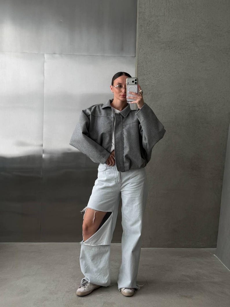 Op de afbeelding: Een grijs kort jasje met kraag en lange mouwen gedragen over een wit shirt, gecombineerd met een lichtblauwe wijde spijkerbroek met grote scheuren bij de knie&euml;n en witte sneakers met een laag model en ronde neuzen.