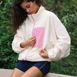 May include: A cream-colored fleece jacket featuring a high collar, a full-length pink front zipper, and a pink chest pocket with a vertical zipper closure. The jacket has long sleeves with elastic cuffs and is paired with dark blue athletic shorts.