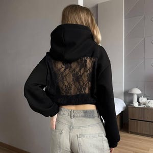 Op de afbeelding: Zwarte hoodie met een kanten achterpaneel. De hoodie heeft lange mouwen en een cropped pasvorm. Het model draagt een lichtblauwe jeans. De achtergrond is een neutrale grijze muur.