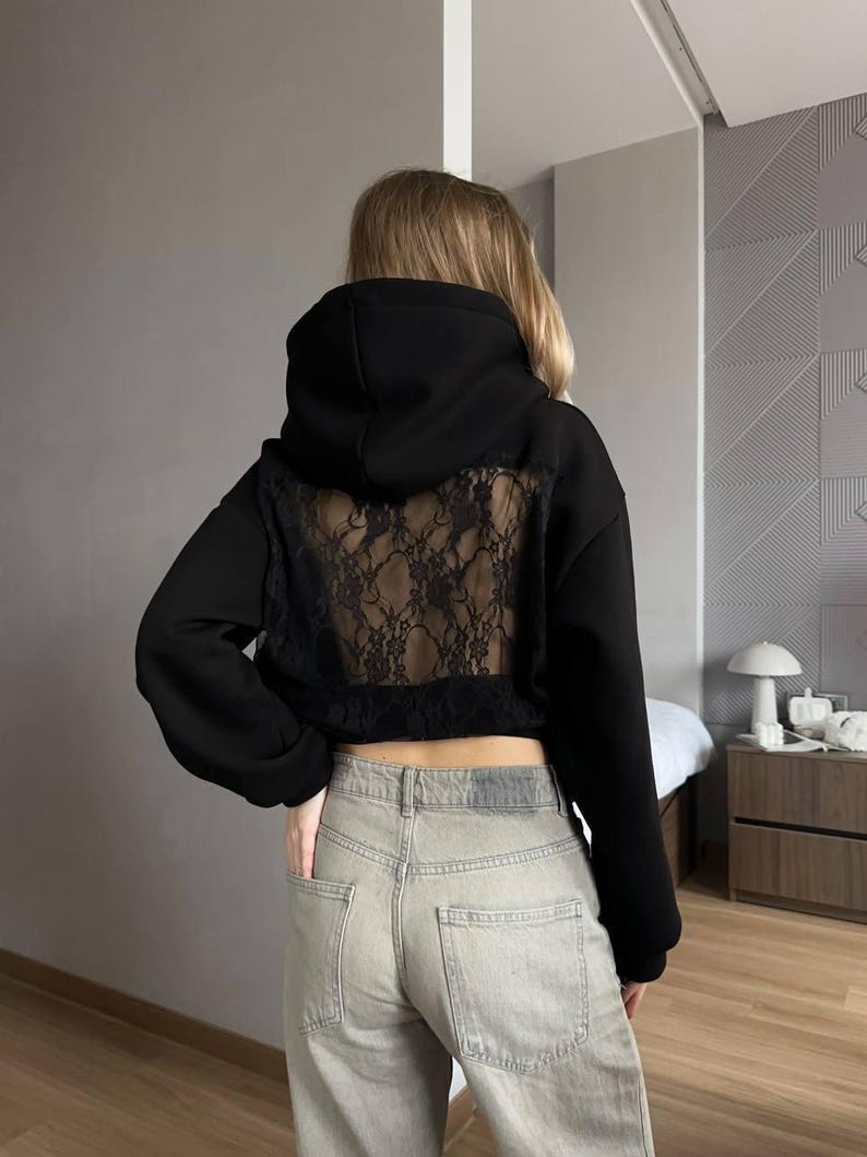 Op de afbeelding: Zwarte hoodie met een kanten achterpaneel. De hoodie heeft lange mouwen en een cropped pasvorm. Het model draagt een lichtblauwe jeans. De achtergrond is een neutrale grijze muur.