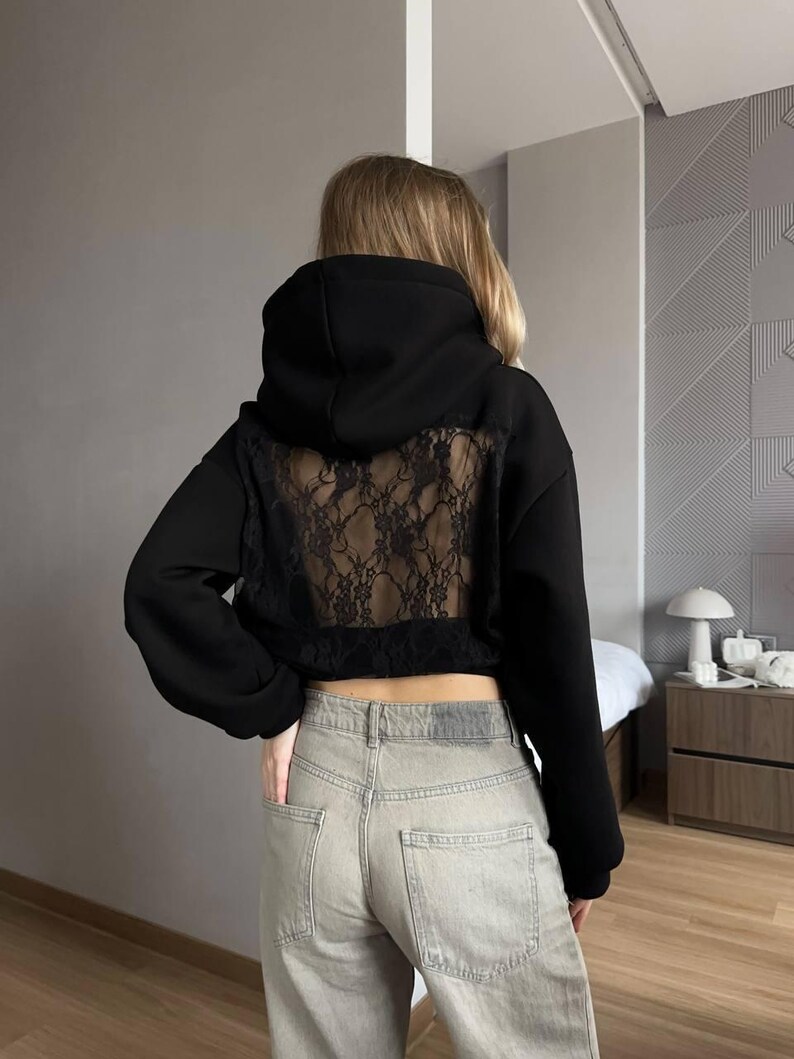 Op de afbeelding: Zwarte hoodie met een kanten achterpaneel. De hoodie heeft lange mouwen en een cropped pasvorm. Het model draagt een lichtblauwe jeans. De achtergrond is een neutrale grijze muur.