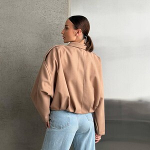 Op de afbeelding: Een beige jas met lange mouwen, een kraag, schouderkleppen en een geplooide zoom, gecombineerd met een lichtblauwe spijkerbroek met een klassiek ontwerp met vijf zakken en een licht versleten afwerking op de broekspijp.