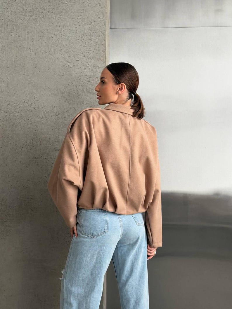 Op de afbeelding: Een beige jas met lange mouwen, een kraag, schouderkleppen en een geplooide zoom, gecombineerd met een lichtblauwe spijkerbroek met een klassiek ontwerp met vijf zakken en een licht versleten afwerking op de broekspijp.