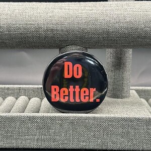 May include: A black, circular button with the words "Do Better" in bright orange lettering. The button is displayed on a grey jewellery display stand. The background is a neutral grey.