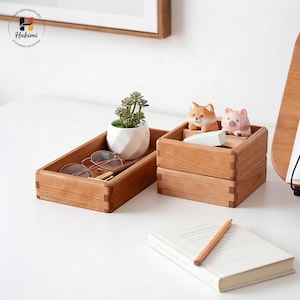 May include: Two wooden rectangular trays, one with eyeglasses, a pen, and a small plant in a white pot. The other tray has a fox and pig figurine. A notebook and pencil are on the table. The trays are made of light brown wood.