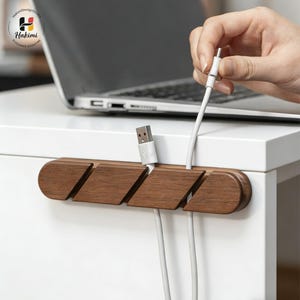 May include: A dark brown wooden cable tidy with angled slots for holding white charging cables. The organiser is mounted on a white surface. A hand is holding a white cable, and a laptop is visible in the background.