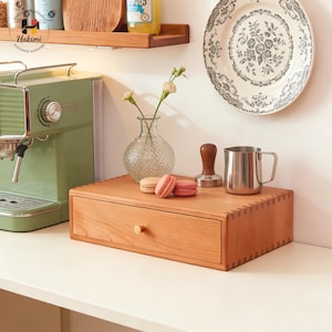 Könnte beinhalten: Holzschublade mit Messingknopf, die eine natürliche Holzmaserung zeigt. Ein Edelstahlbecher, ein Holz-Tamper und eine Glasvase mit Blumen sind ebenfalls vorhanden. Macarons setzen farbliche Akzente.