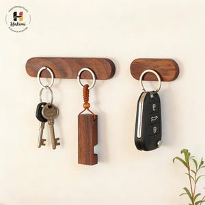 May include: Two wooden key holders with silver key rings. One holds keys, the other a wooden bottle opener, and the third a car key. The key holders are made of dark wood and mounted on a white wall.