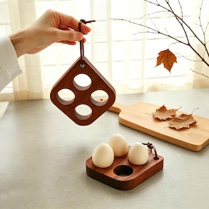May include: Two square dark wood egg holders with four circular cutouts each. One holder is suspended by a brown leather strap, while the other rests horizontally, containing three smooth, off-white eggs in its circular openings.