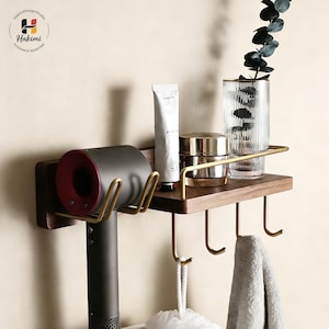 Puede incluir: Estante de baño de madera con detalles en latón. Cuenta con un soporte para secador de pelo, ganchos para toallas y un estante que contiene productos para el cuidado de la piel y un jarrón decorativo. El estante está montado en la pared.