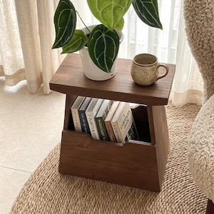May include: A dark brown wooden side table with a flat top and a lower storage compartment. The top holds a white potted plant with large green leaves and a speckled beige ceramic mug. Several books are stored upright in the lower section.