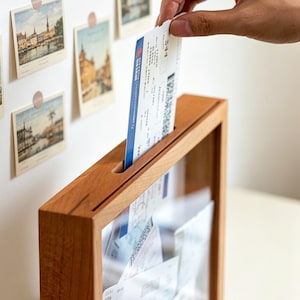 Puede incluir: Un marco de madera rectangular tipo caja con un panel frontal transparente y una ranura en el borde superior. Un billete de papel blanco con detalles azules se está insertando en la ranura, mientras otros billetes similares se ven dentro.