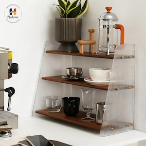 Pode incluir: Uma estante de madeira de três níveis com painéis laterais transparentes e canelados. As prateleiras exibem uma variedade de itens, incluindo uma planta em vaso, xícaras de café, uma prensa francesa e artigos de vidro. A unidade é colocada contra uma parede branca.