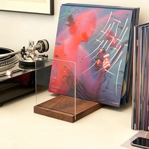 May include: A wooden and clear acrylic record holder displaying a vinyl album with a colorful cover. The album art features a person with red and blue tones. The holder is on a white surface, with a turntable in the background.