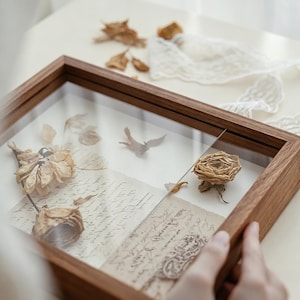 Puede incluir: Un marco de madera rectangular tipo vitrina que contiene rosas secas prensadas y documentos de papel pergamino con escritura a mano de estilo antiguo. El marco cuenta con un frente de vidrio transparente y un acabado de madera natural.
