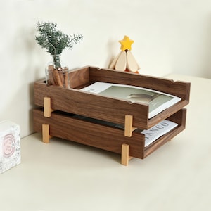K&ouml;nnte beinhalten: Ein zweistufiger Schreibtisch-Organizer aus Holz mit dunkelbraunem Finish. Der Organizer hat eine rechteckige Form mit einem Tablett oben und einem zweiten Tablett darunter. Die Tabletts werden von hellfarbigen Holzst&uuml;tzen getragen. Oben steht eine kleine Glasvase mit Gr&uuml;nzeug.