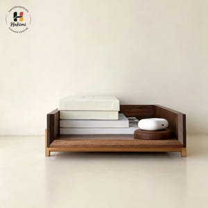 K&ouml;nnte beinhalten: Ein h&ouml;lzerner Schreibtisch-Organizer mit rechteckiger Form, der einen Stapel B&uuml;cher und wei&szlig;e Boxen enth&auml;lt. Ein kleines, wei&szlig;es, oval geformtes Objekt liegt auf einem runden, braunen Holzsockel. Der Organizer ist aus dunkelbraunem Holz gefertigt.