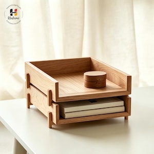 K&ouml;nnte beinhalten: Ein helles Schreibtisch-Organizer aus Holz mit zwei gestapelten Ablagen. Ein kleiner, runder Holzbeh&auml;lter befindet sich auf der oberen Ablage. Der Organizer enth&auml;lt zwei B&uuml;cher. Das Logo "Hakimi Design Studio" befindet sich oben links.